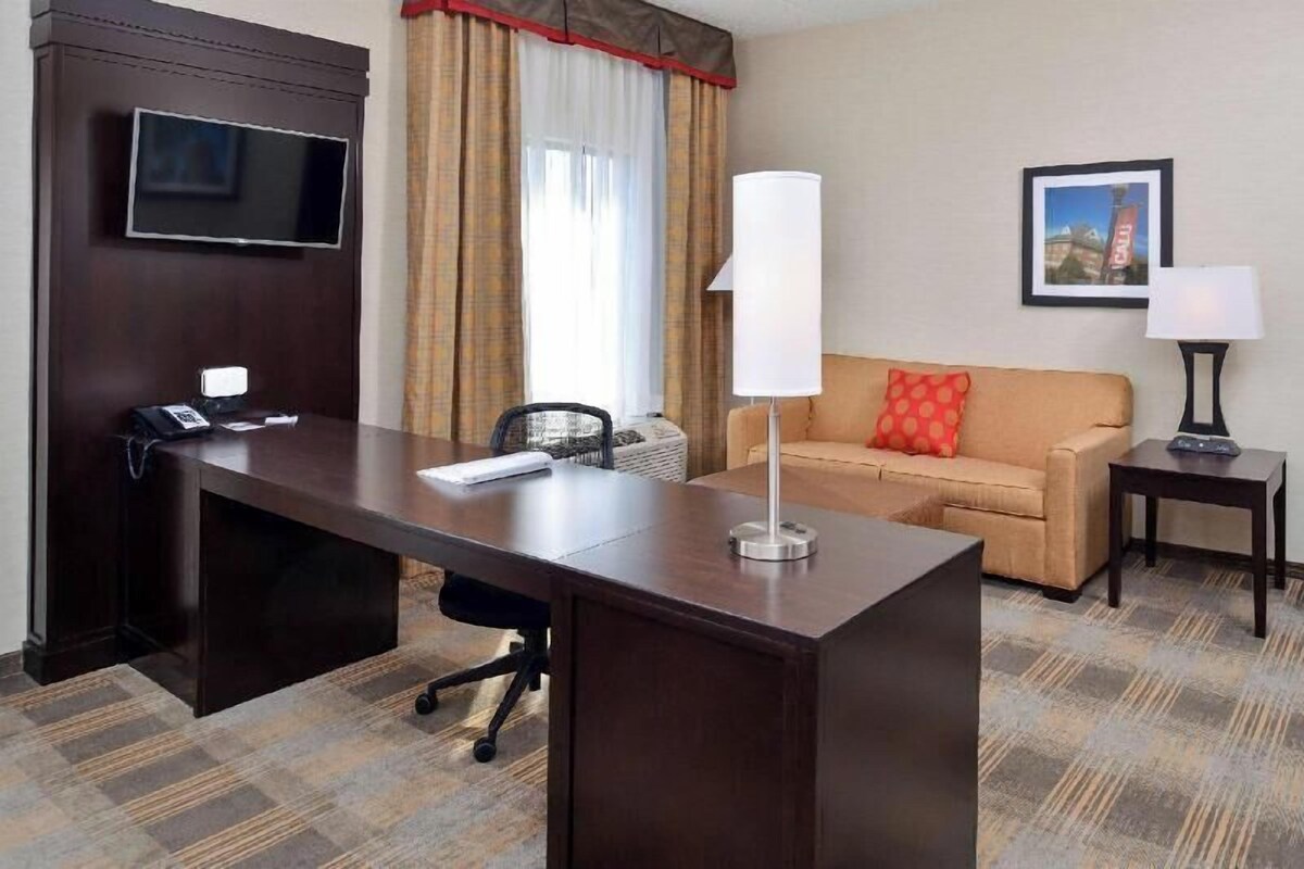 A hotel room with a desk, a television, a sofa, a lamp, and a framed picture on the wall.