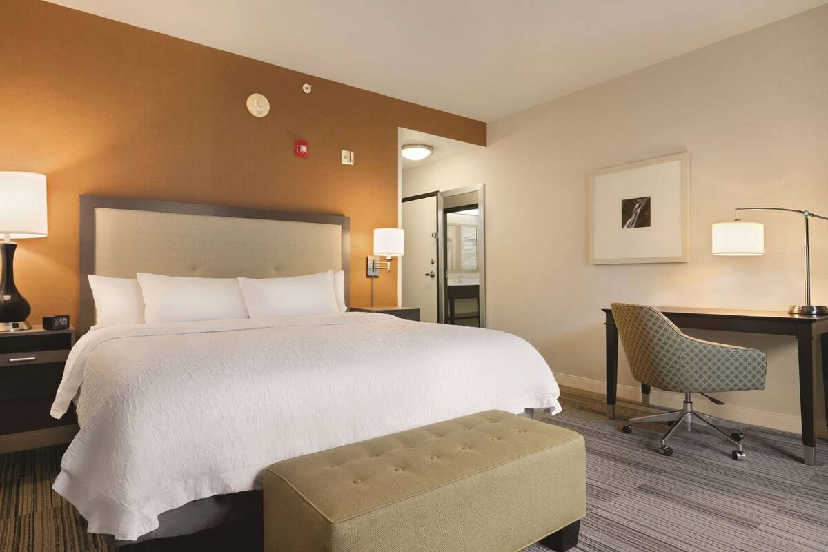 A hotel room with a large bed, a desk, a chair, a lamp, and a small ottoman.
