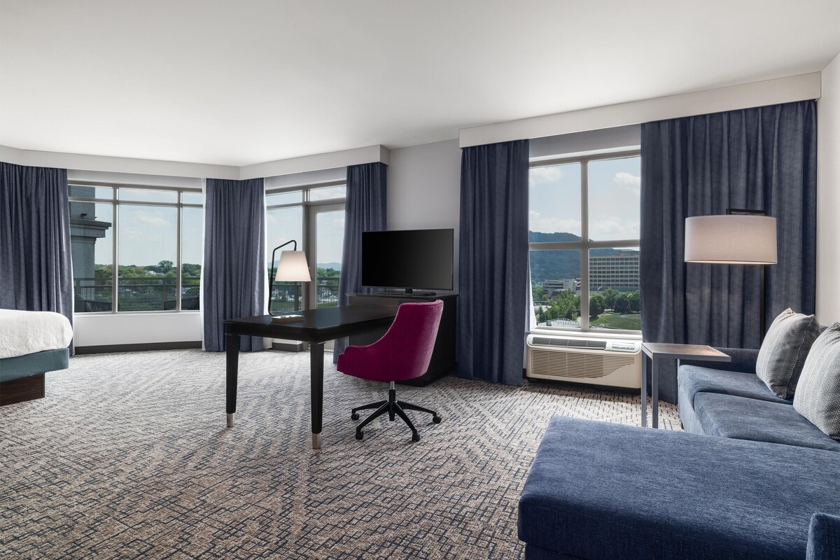 Una habitación de hotel con una cama, un escritorio con silla, un televisor, un sofá y ventanas amplias con cortinas.