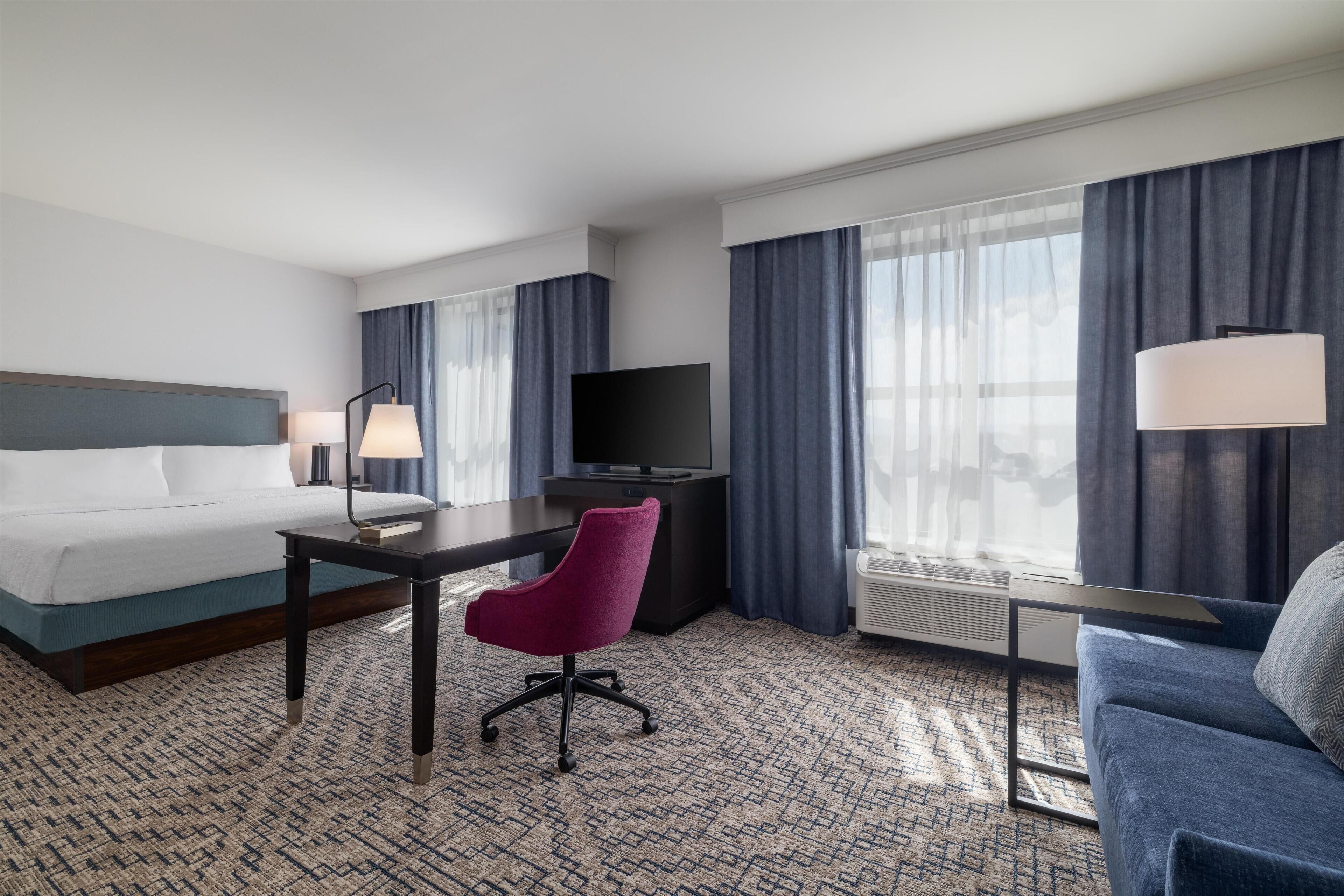 A hotel room with a bed, desk, chair, TV, and large windows with curtains.