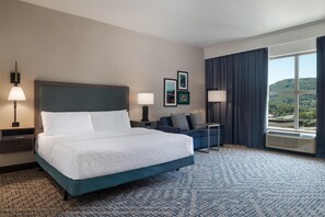 A hotel room with a large bed, two bedside tables with lamps, a small desk, a sofa, and a large window with a view of the outdoors. - Hampton Inn & Suites Roanoke-Downtown (Roanoke)