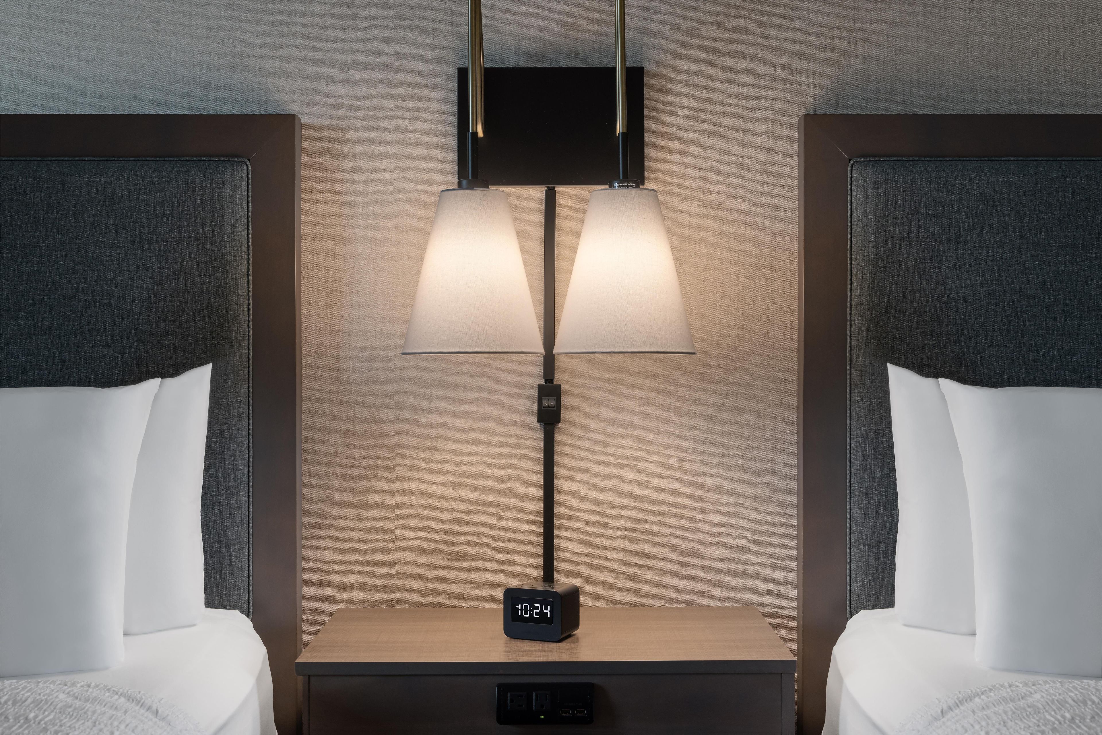 A hotel room with two beds, a nightstand with a digital clock, and a wall-mounted lamp.
