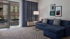 Una habitación de hotel moderna con un sofá azul, una mesita, una lámpara de pie y un ventanal con vistas a un edificio.