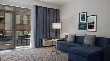 A modern hotel room with a blue sofa, a small table, a floor lamp, and a large window with a view of a building.