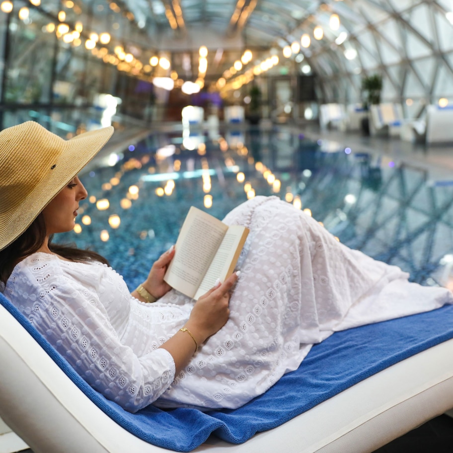 A person lying on a lounge chair reading a book in a modern indoor setting with glass flooring and decorative lighting.