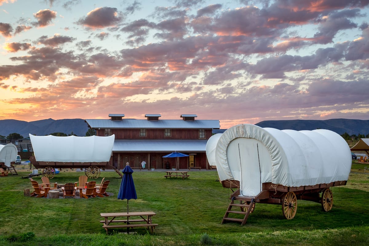 A covered wagon and a rustic building with a wooden exterior and a flat roof, set against a backdrop of mountains and a sunset sky.