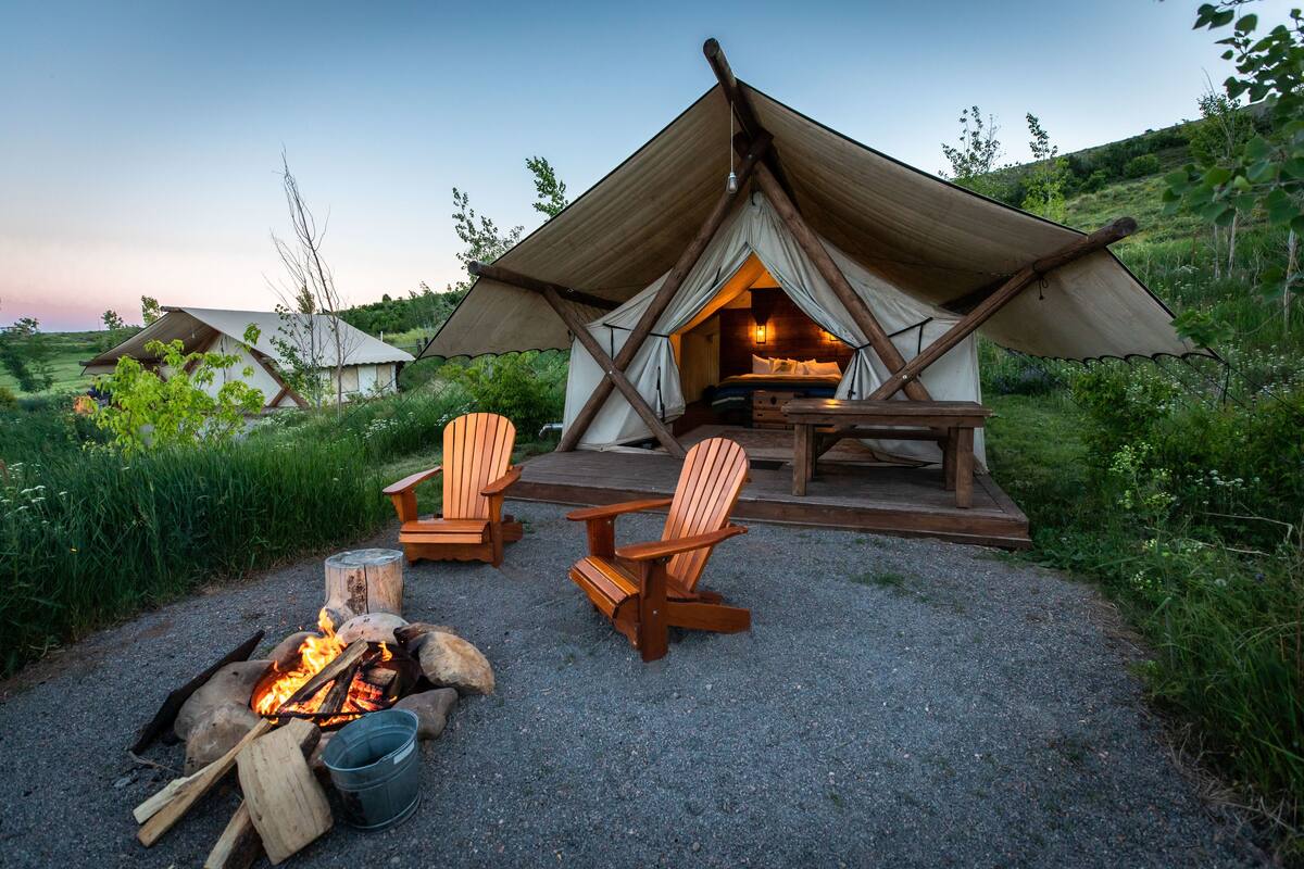 A rustic tent-style accommodation with a fire pit, wooden chairs, and a bed inside.