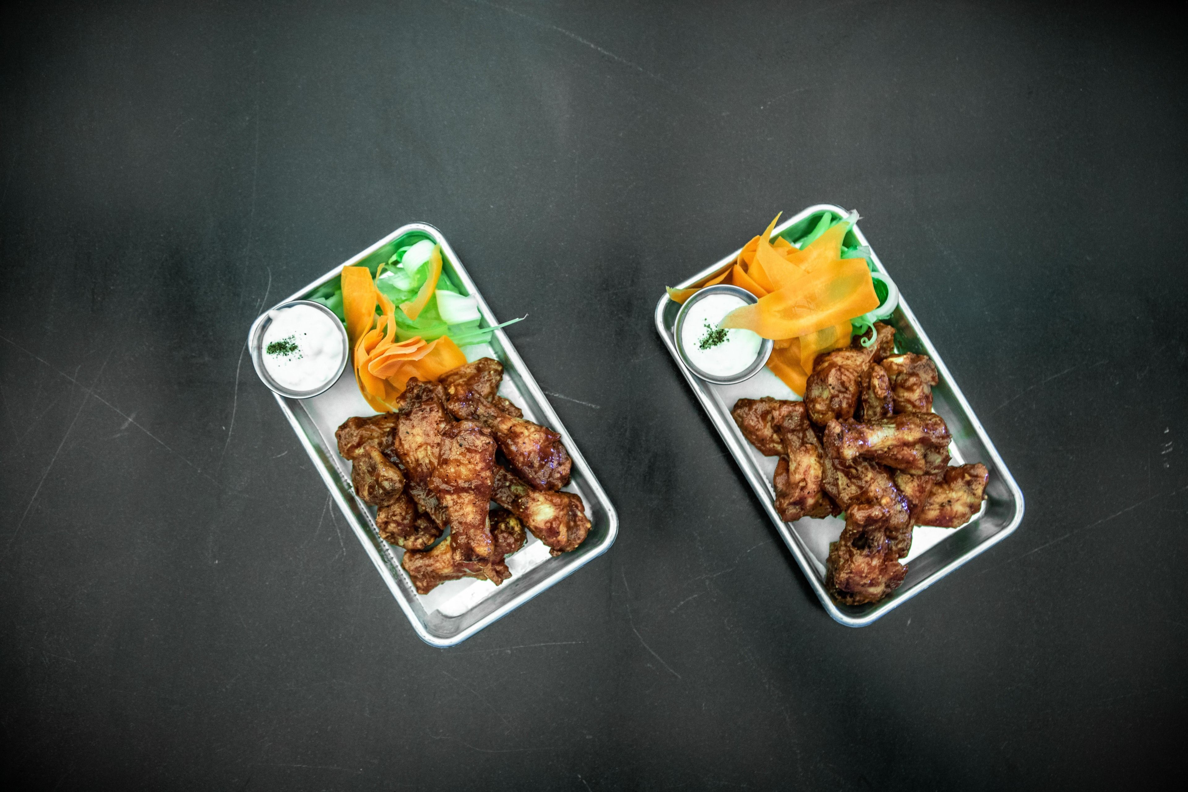 Dos bandejas de metal con alitas de pollo a la parrilla, pimientos amarillos como acompañamiento y un recipiente pequeño con salsa.