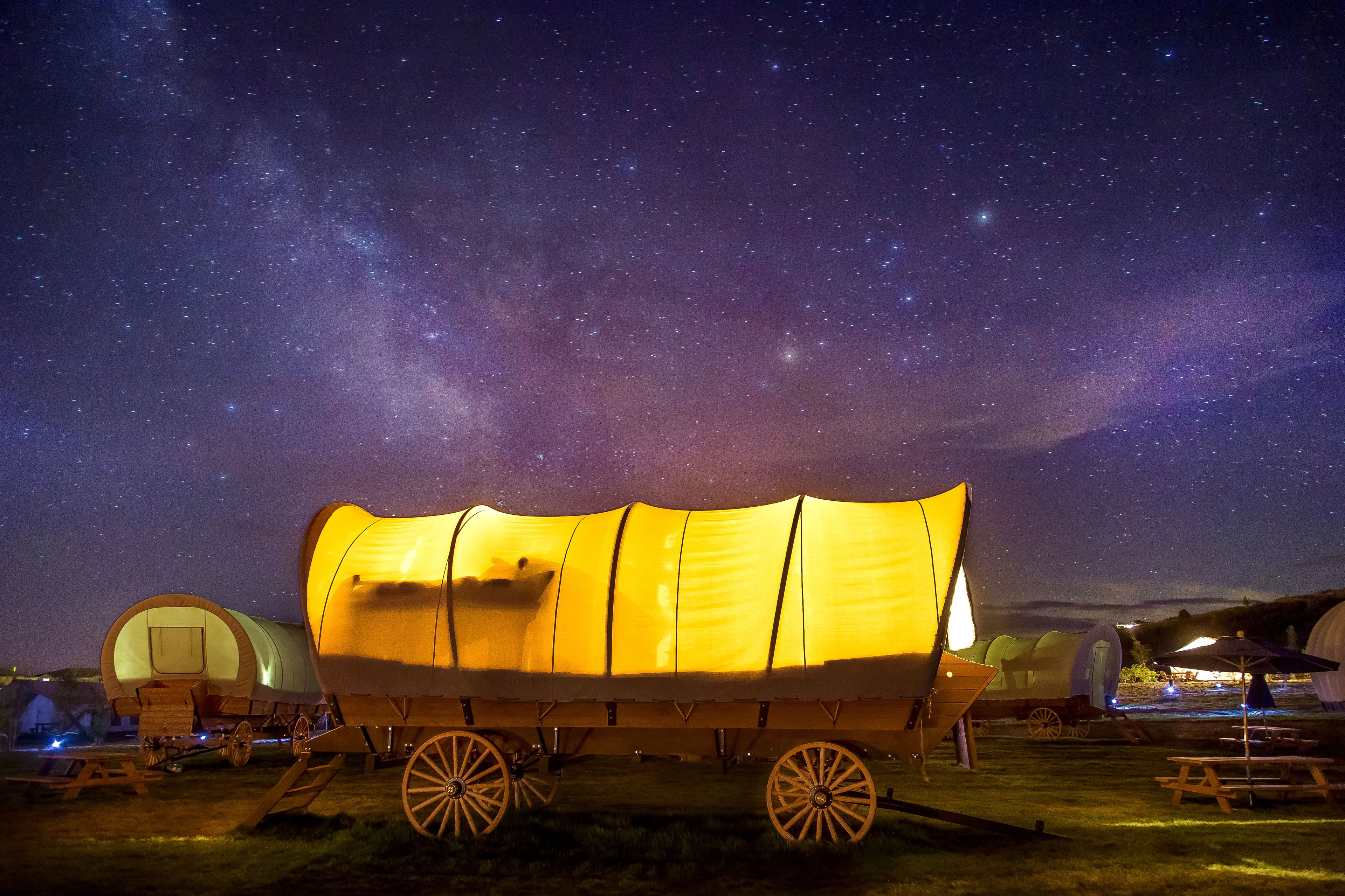 A covered wagon under a starry night sky, surrounded by tents and picnic tables.