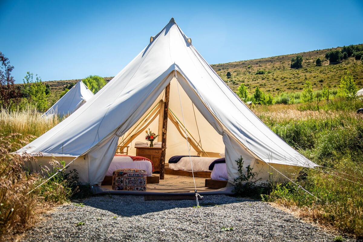 A white tent with two beds and a small table, set in a grassy area with a hill in the background.