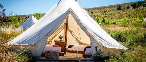 A white tent with two beds and a small table, set in a grassy area with a hill in the background.
