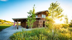 A rustic wooden cabin with a covered porch, surrounded by a natural landscape with wildflowers and trees. - Conestoga Ranch (Garden City)