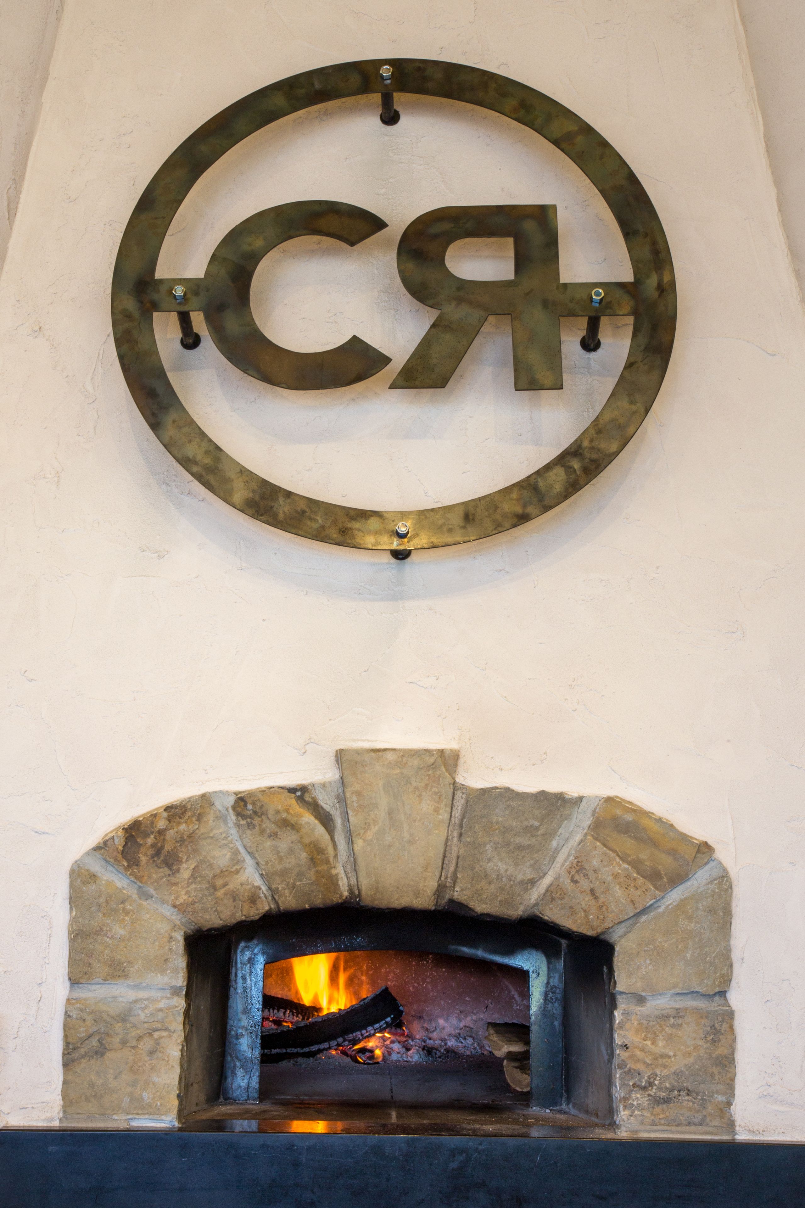 A rustic stone fireplace with a fire burning inside, a metal sign with Cyrillic letters above it, and a stone archway framing the fireplace.
