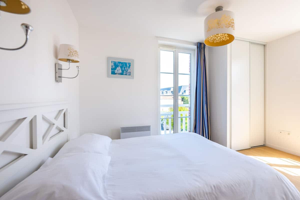 Une chambre avec un grand lit, une fenêtre avec des rideaux, une lampe fixée au mur et un petit tableau.