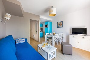 A modern living room with a blue sofa, a white coffee table, a grey ottoman, a dining area with a white table and chairs, a kitchen with blue cabinets, and a TV on a white cabinet. - Odalys Résidence Les Dunettes (Cabourg)