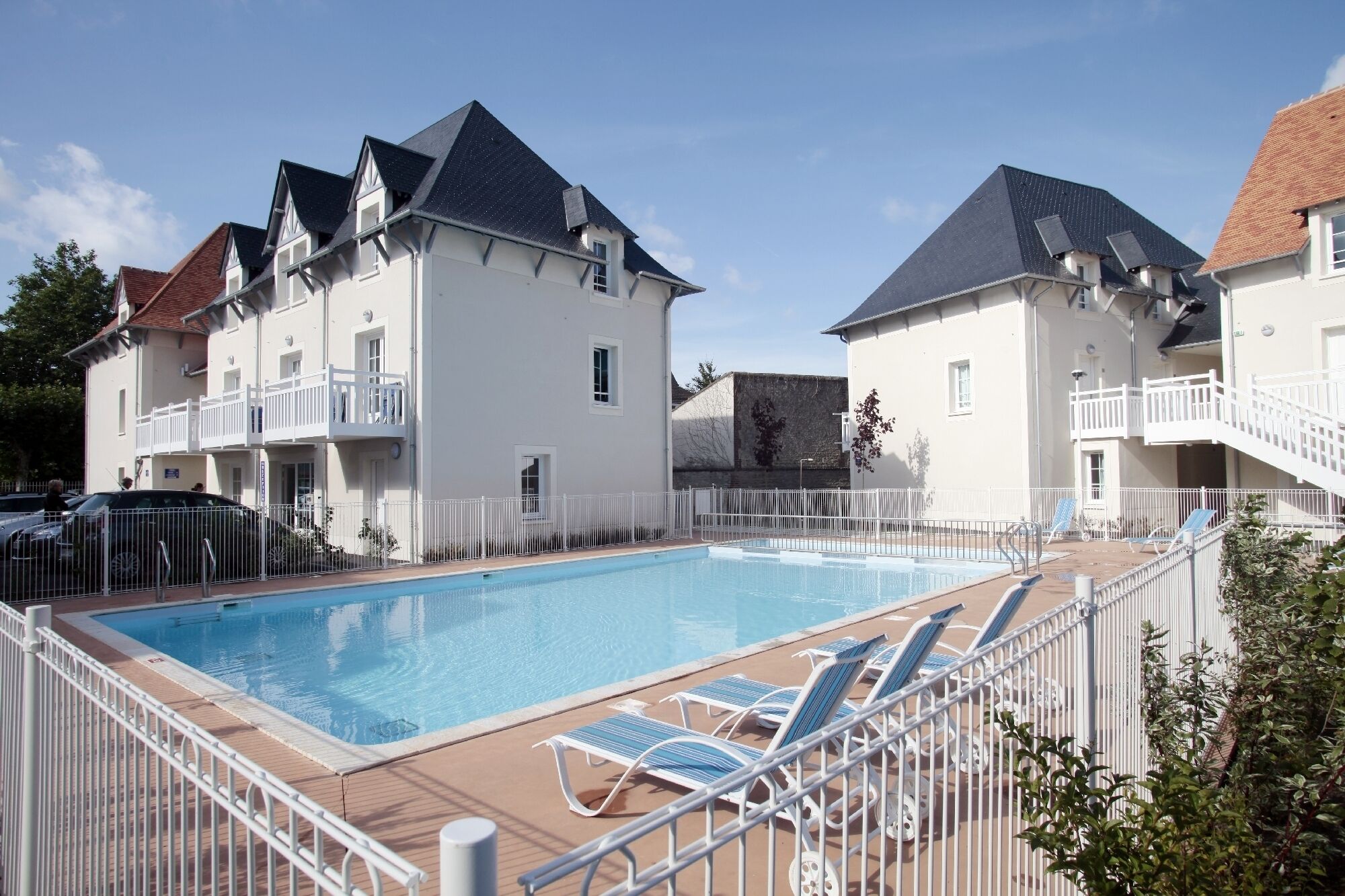 Ein Schwimmbecken mit Liegestühlen, umgeben von einem weißen Zaun, neben einem Gebäude mit einem Balkon.