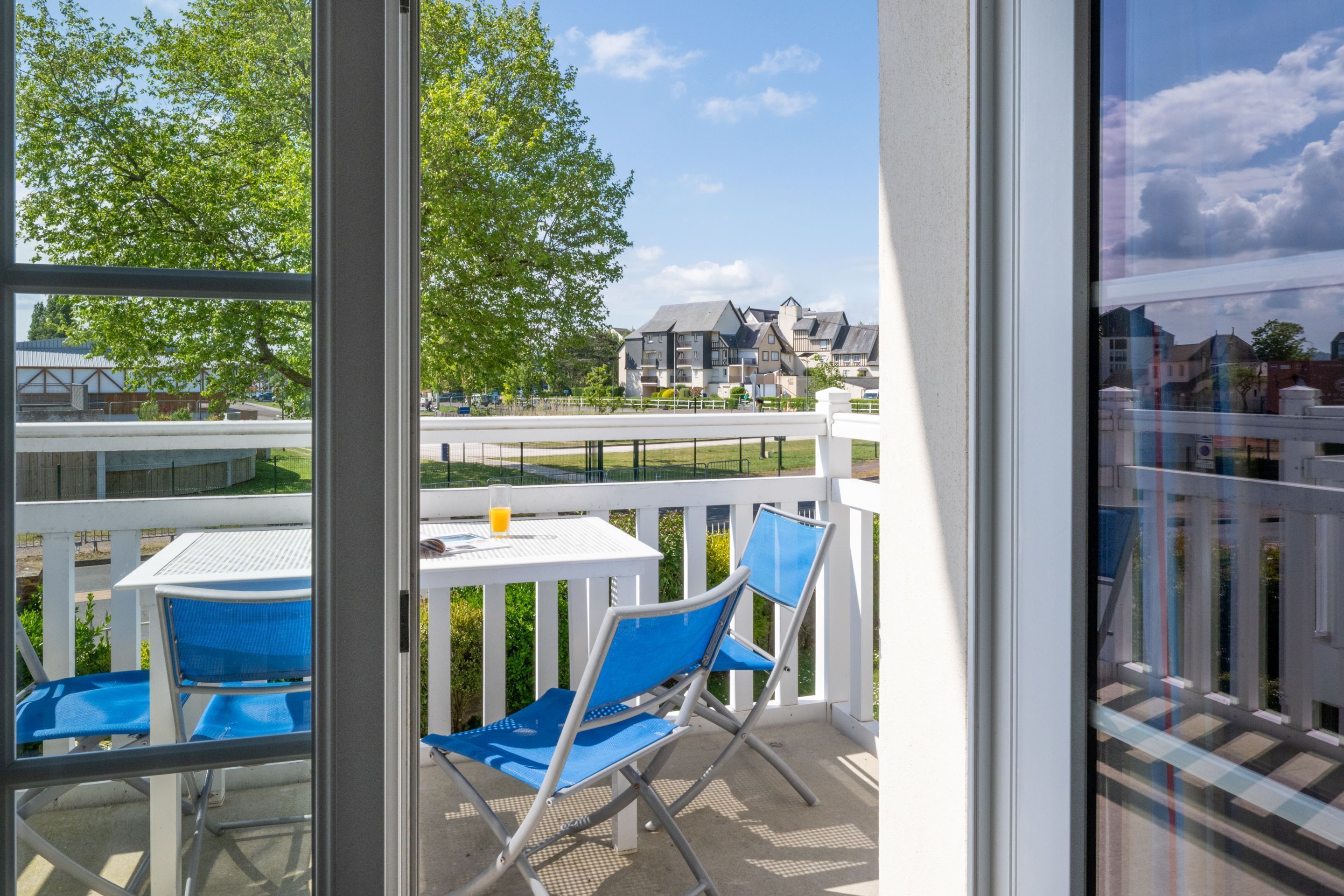 En balkon med blå stole og et hvidt bord, med udsigt over et boligområde med huse og en klar himmel.