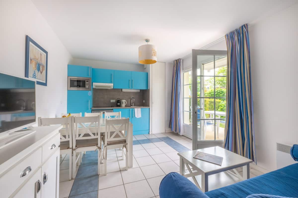 Une cuisine moderne avec des meubles bleus, un coin repas avec des chaises blanches et une grande fenêtre agrémentée de rideaux.