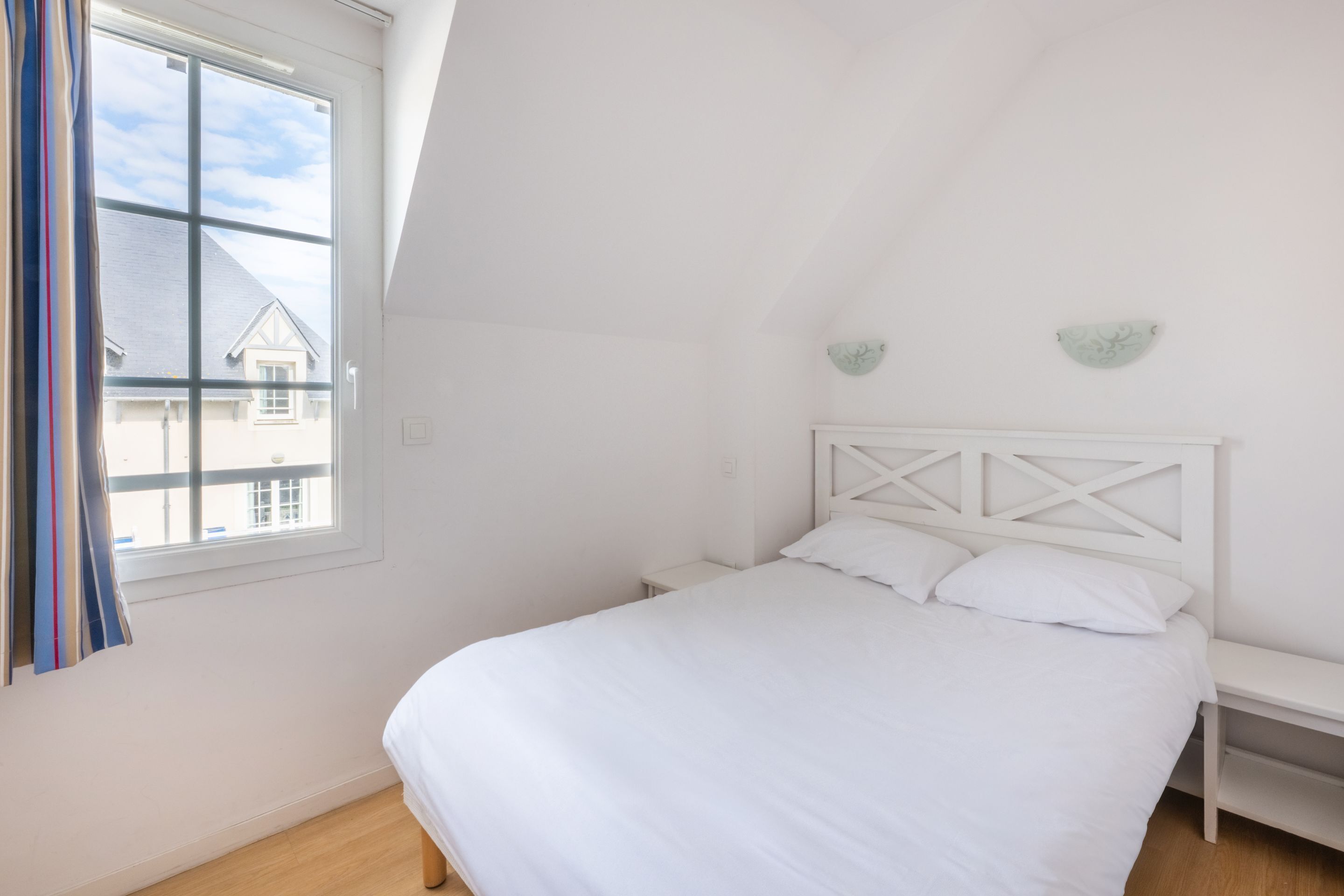 Ein Schlafzimmer mit einem weißen Bett, einem Nachttisch, einem Fenster mit Vorhängen und Blick auf ein Haus.