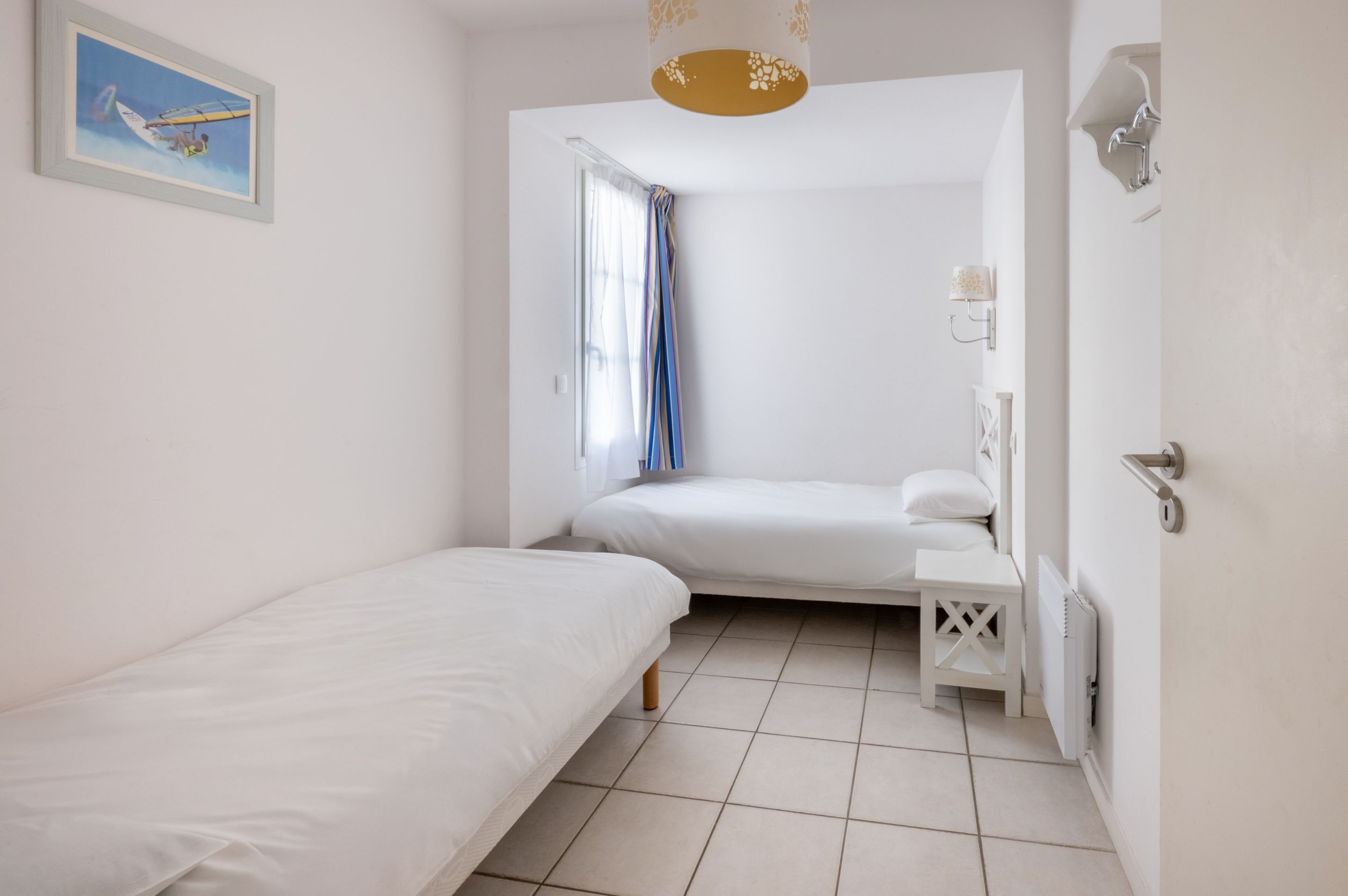 A small, clean room with a bed, a small table, a nightstand, a wall-mounted lamp, and a window with curtains.