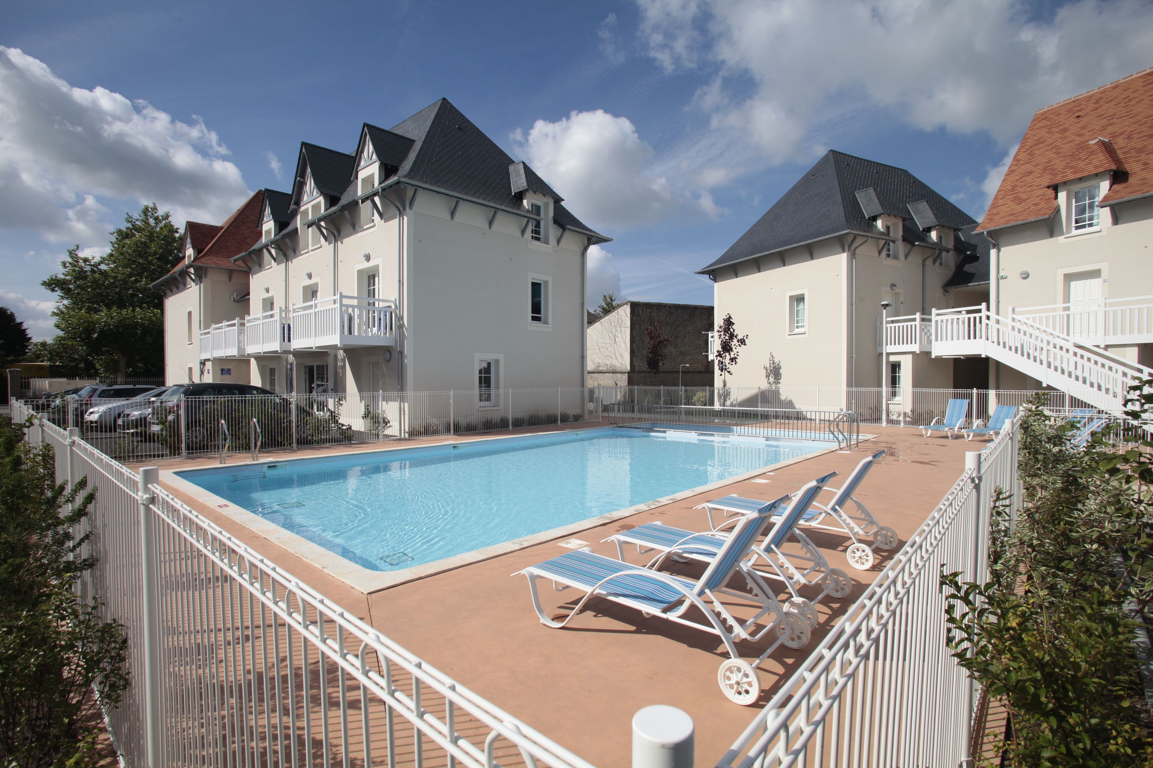 A swimming pool with lounge chairs, surrounded by a white fence, and adjacent to a row of houses with balconies.