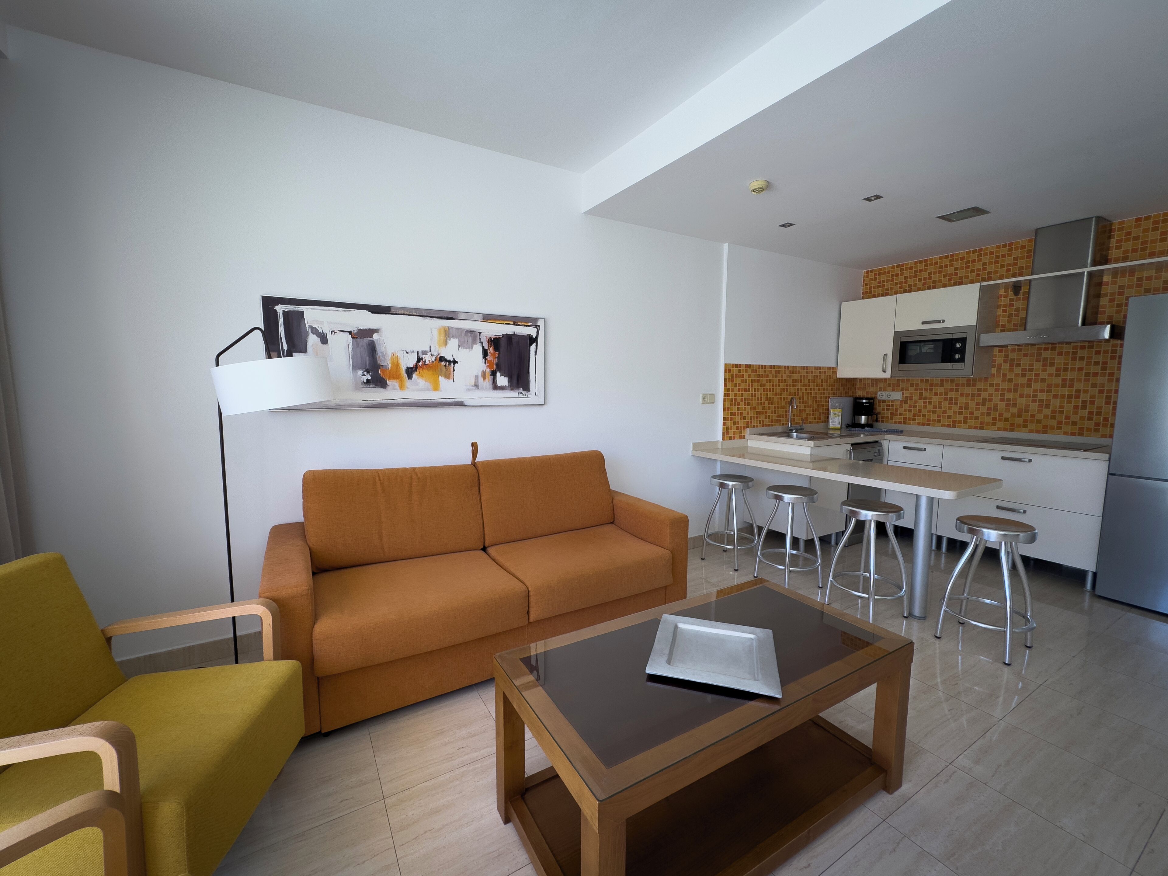 A modern living room with a sofa, a coffee table, and a kitchen area with a dining table and stools.