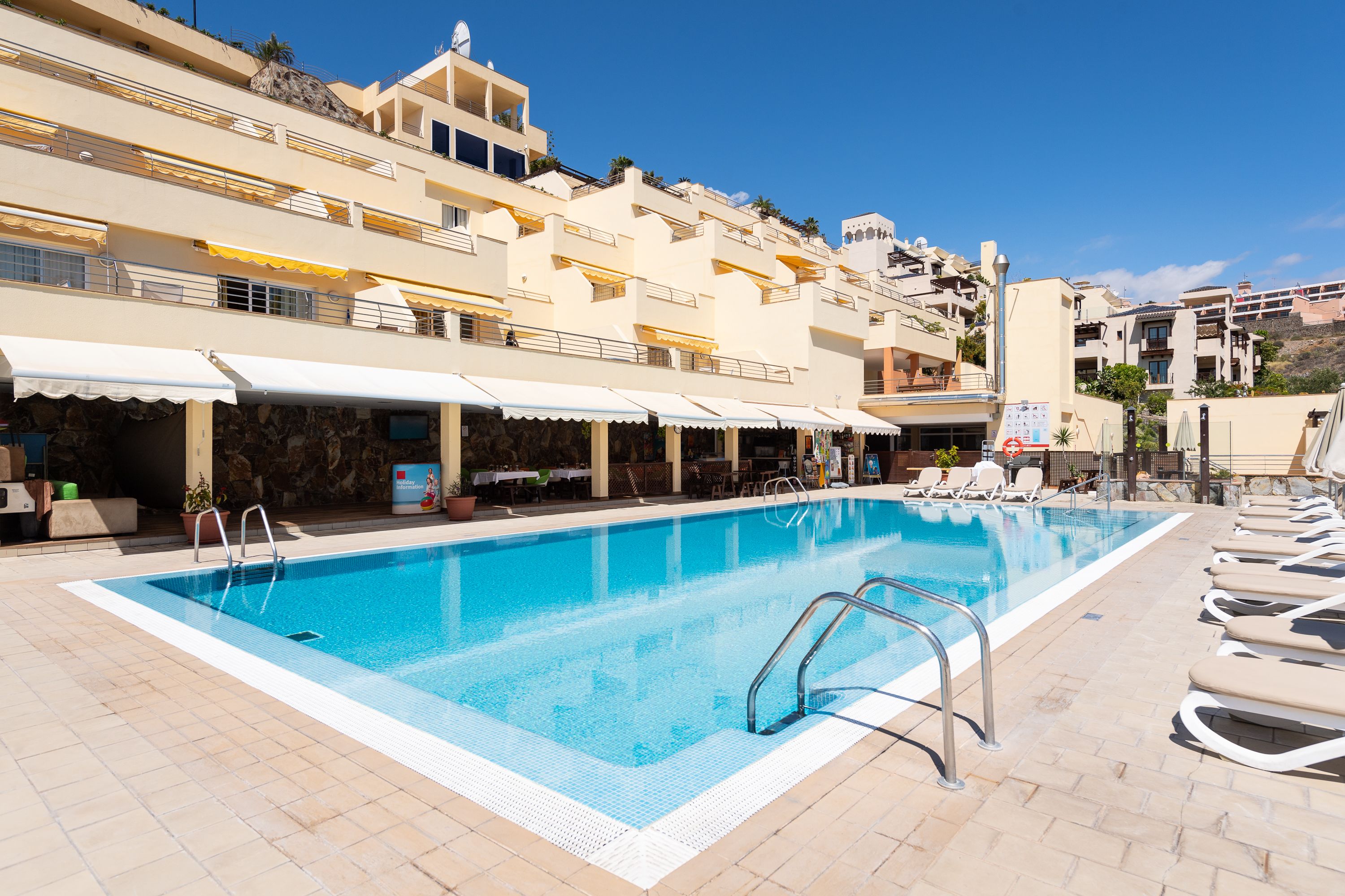 Uma piscina com espreguiçadeiras e guarda-sóis, cercada por edifícios altos com varandas e áreas de estar ao ar livre.