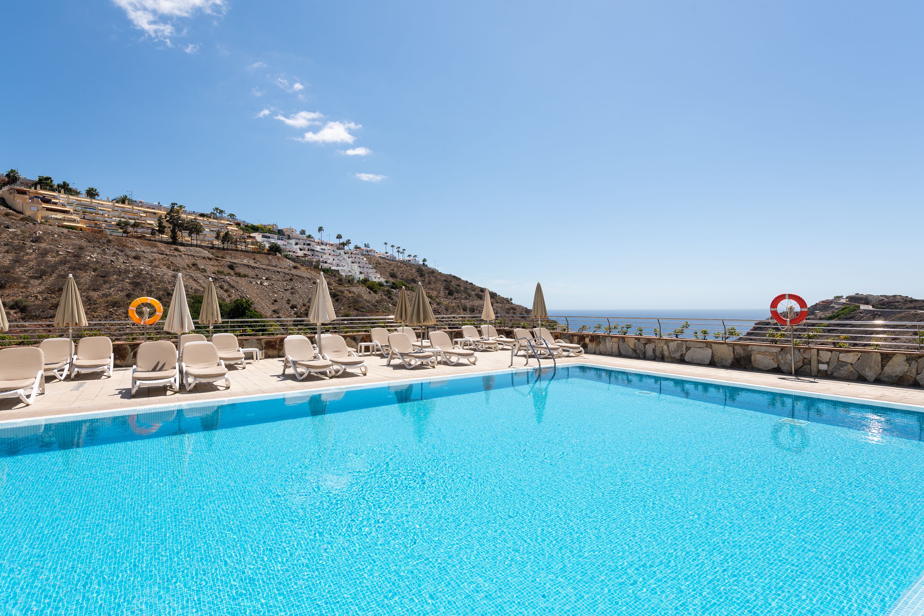 A swimming pool with lounge chairs and umbrellas, overlooking a hillside with buildings and a view of the sea.