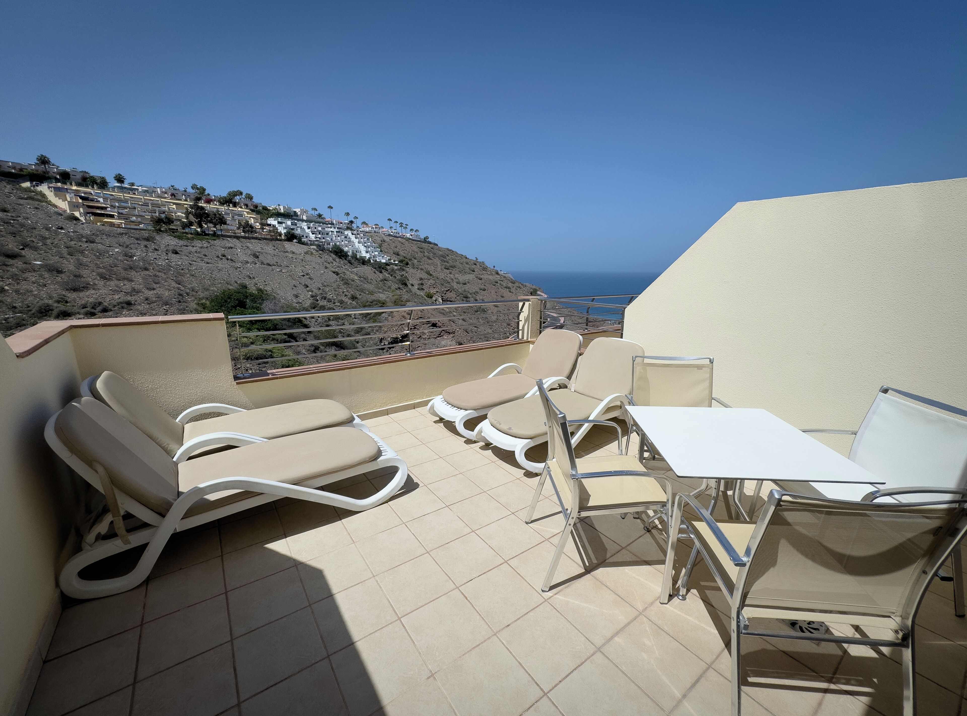 Une terrasse avec des chaises longues beiges et une table, donnant sur une colline parsemée de bâtiments et sous un ciel bleu dégagé.