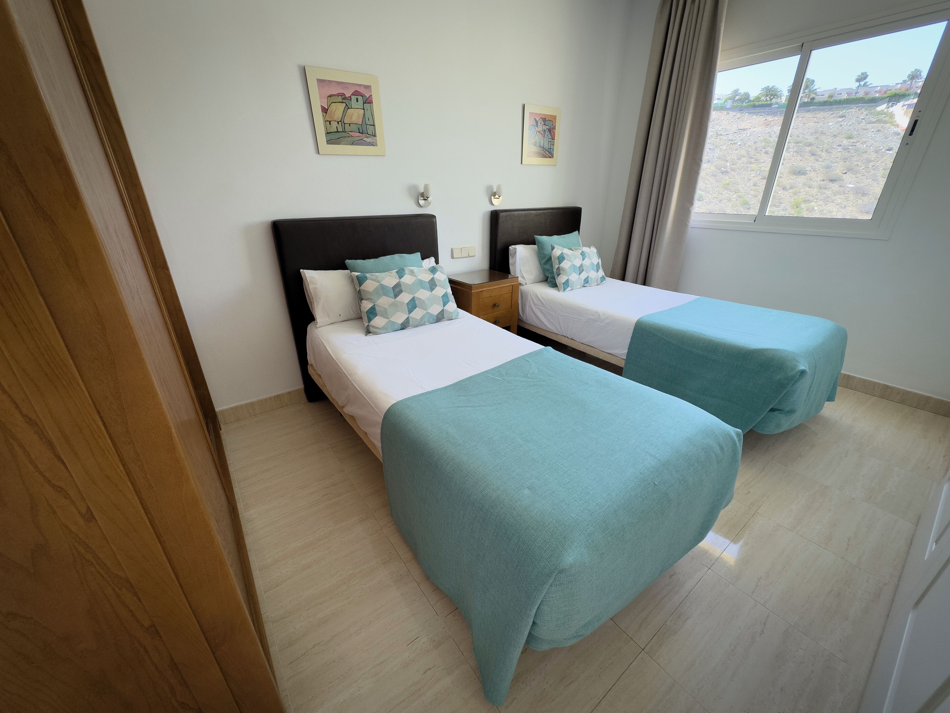 A hotel room with two beds, each with a teal bedspread and matching pillows. There are two wall-mounted paintings, a wooden nightstand with a lamp, and a window with curtains.