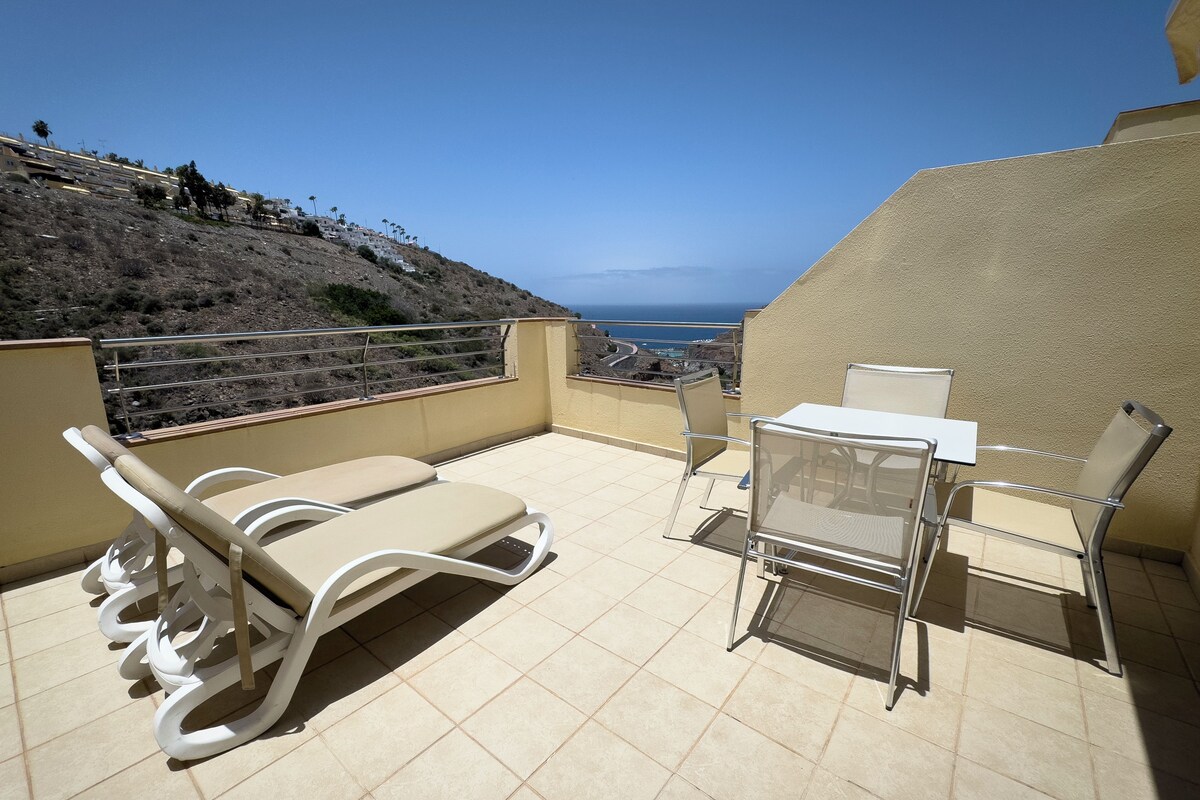 A balcony with a table and chairs, a white cushioned lounge chair, and a view of the sea and hills.