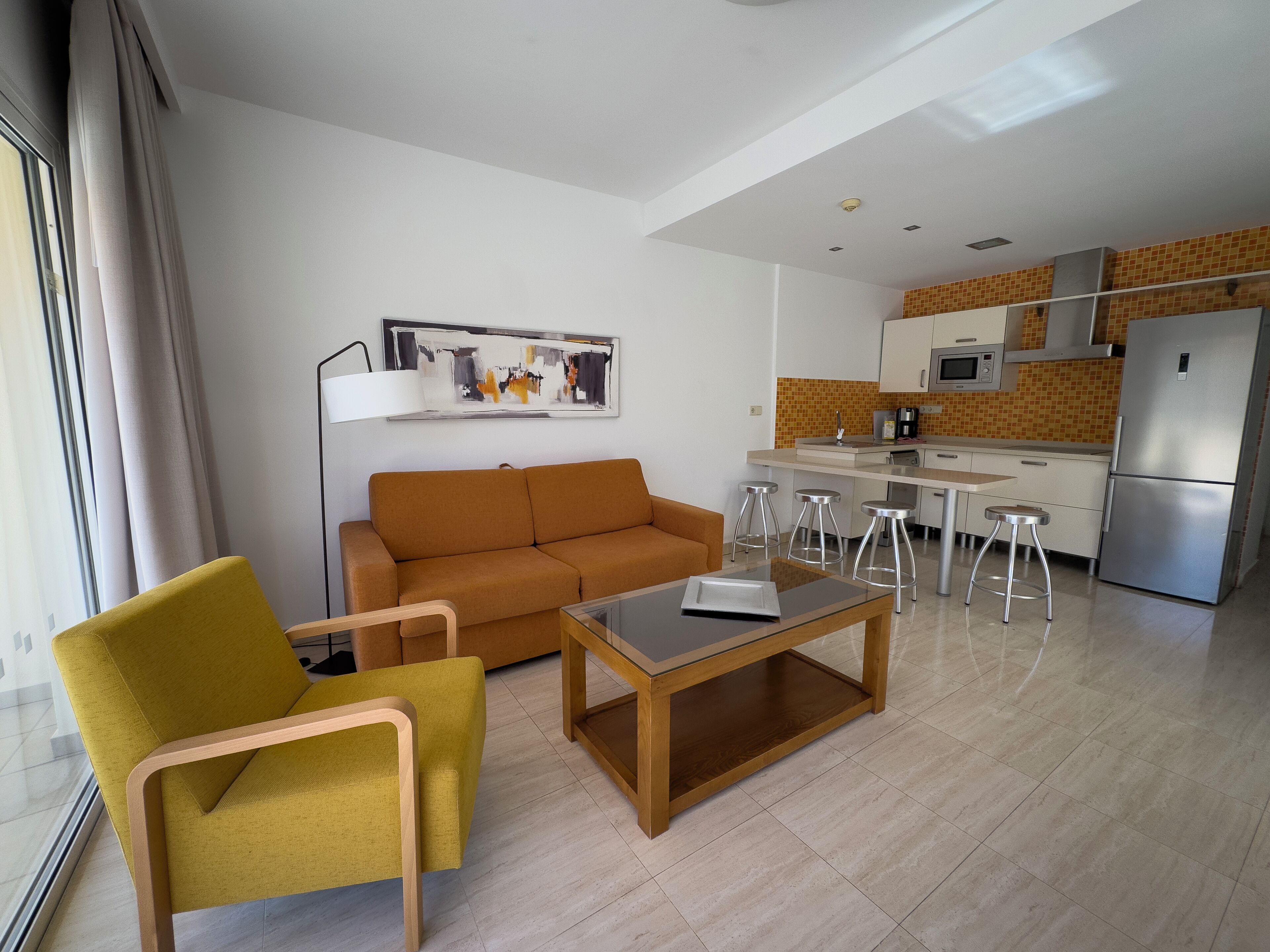 A modern living room with a sofa, a yellow armchair, and a coffee table. A kitchen with a refrigerator, stove, and stools is visible in the background.