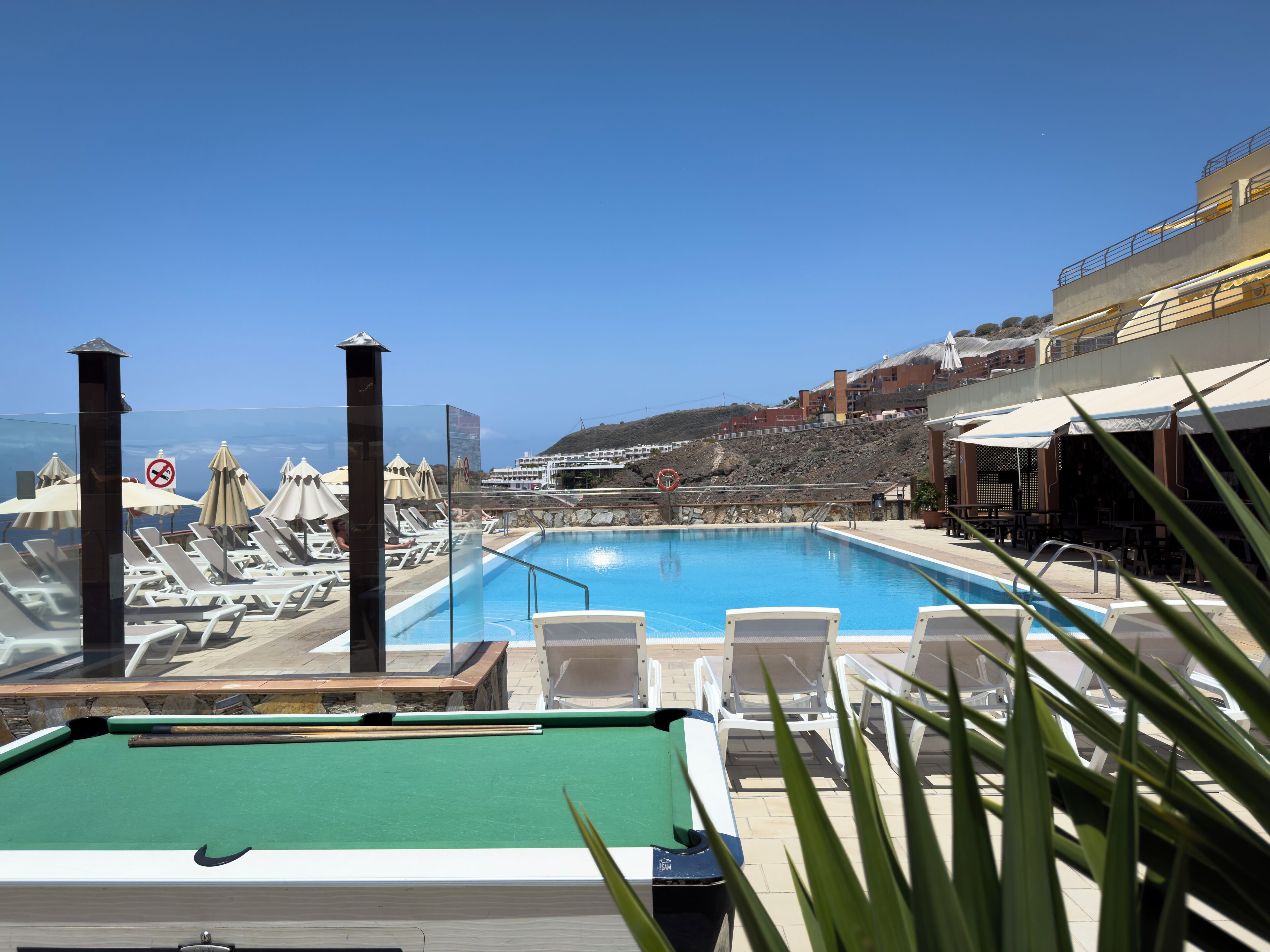 A pool area with lounge chairs, a pool table, and a view of the sea.