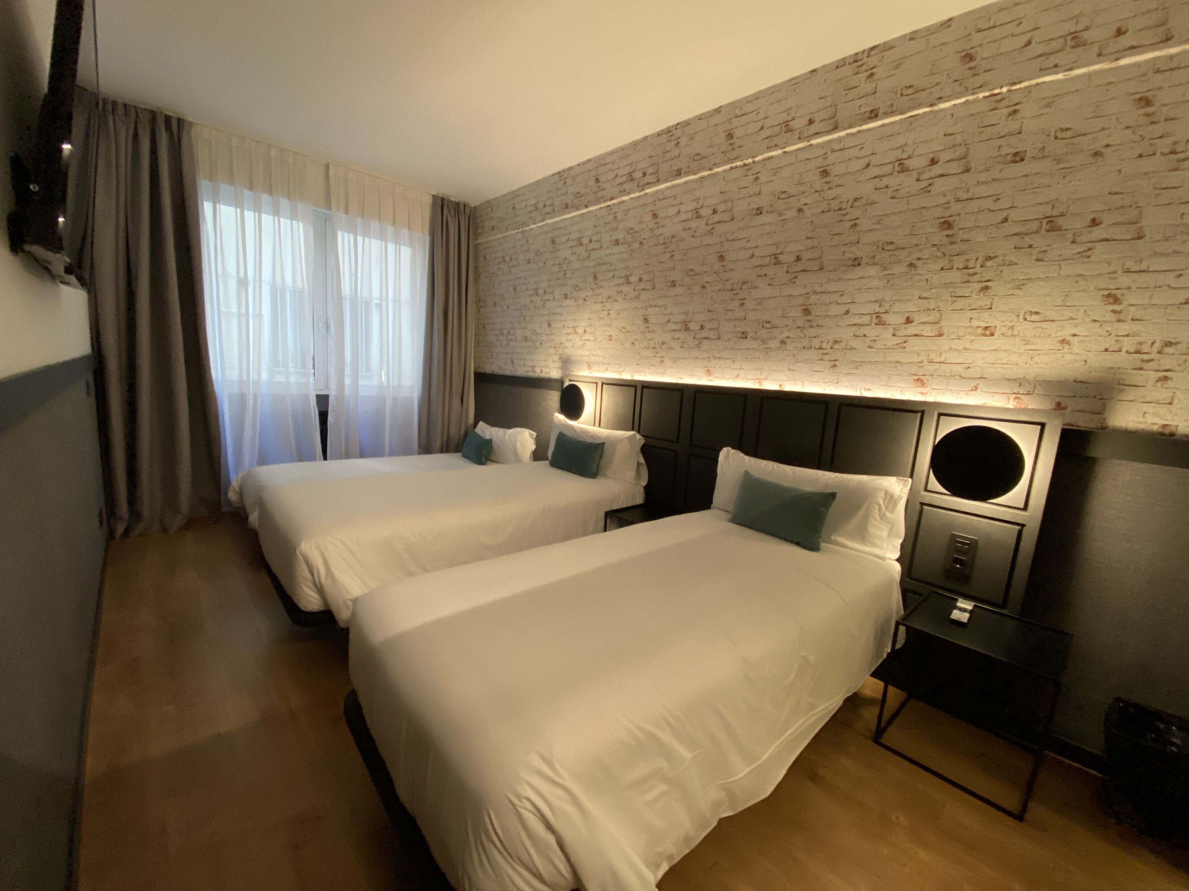 Habitación de hotel con dos camas, una pared de ladrillos, un televisor montado y una ventana con cortinas.