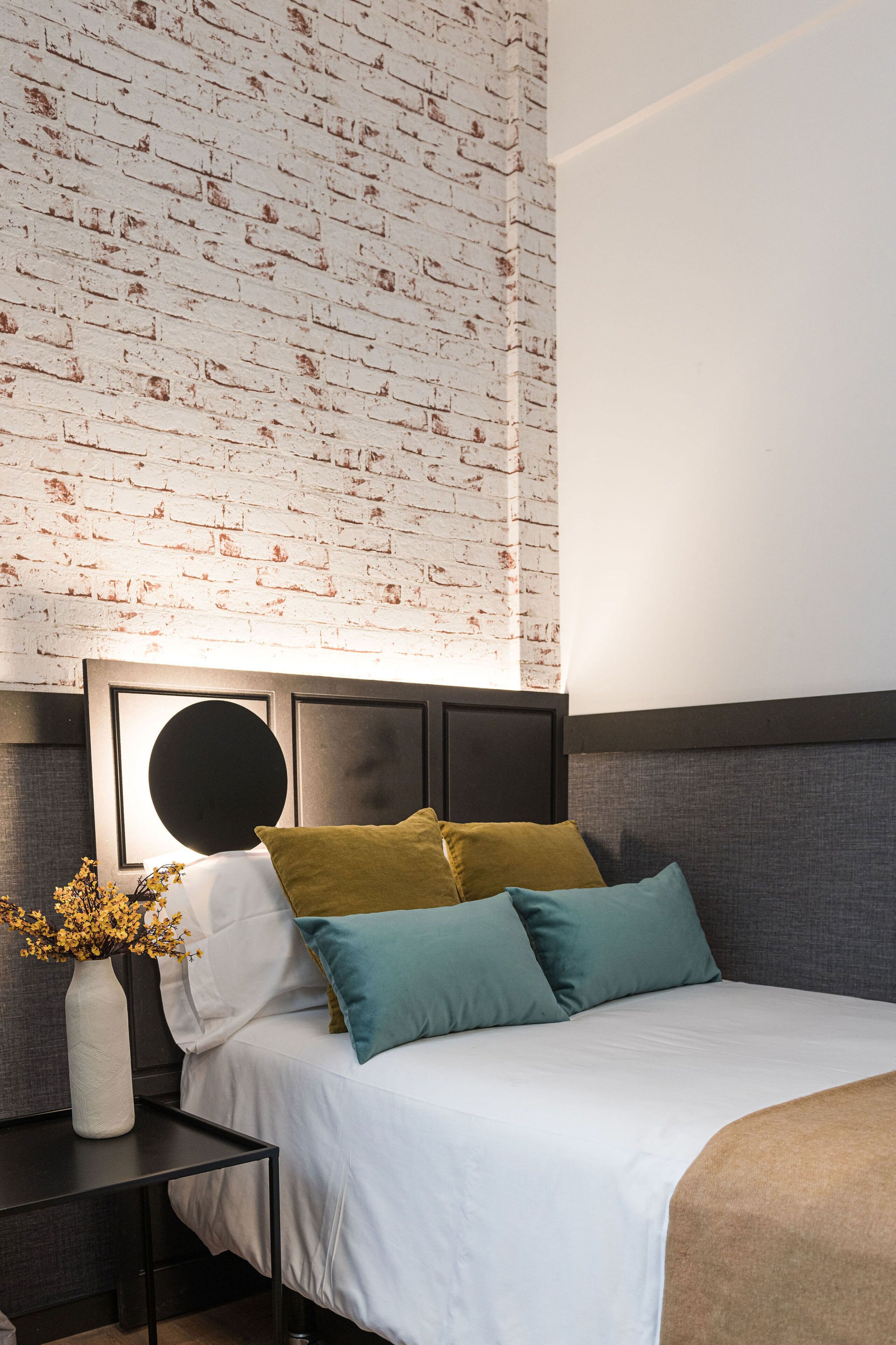 Un dormitorio ordenado con pared de ladrillo, una cama con ropa blanca y varios almohadones, una mesita de noche con un florero y un cabecero moderno.