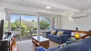 Ein geräumiges Wohnzimmer mit einem blauen Sofa, einem Couchtisch und einem Fernseher. Ein Balkon bietet Blick auf grüne Natur.