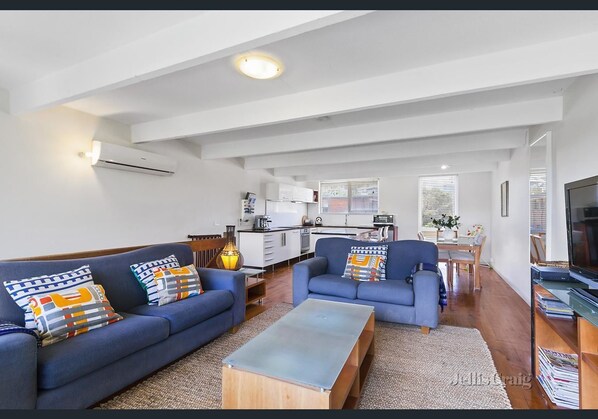 A modern living room with two sofas, a coffee table, and a TV. There is a kitchen in the background with white cabinets and a dining area. - Bayside St Johns Close (Blairgowrie)