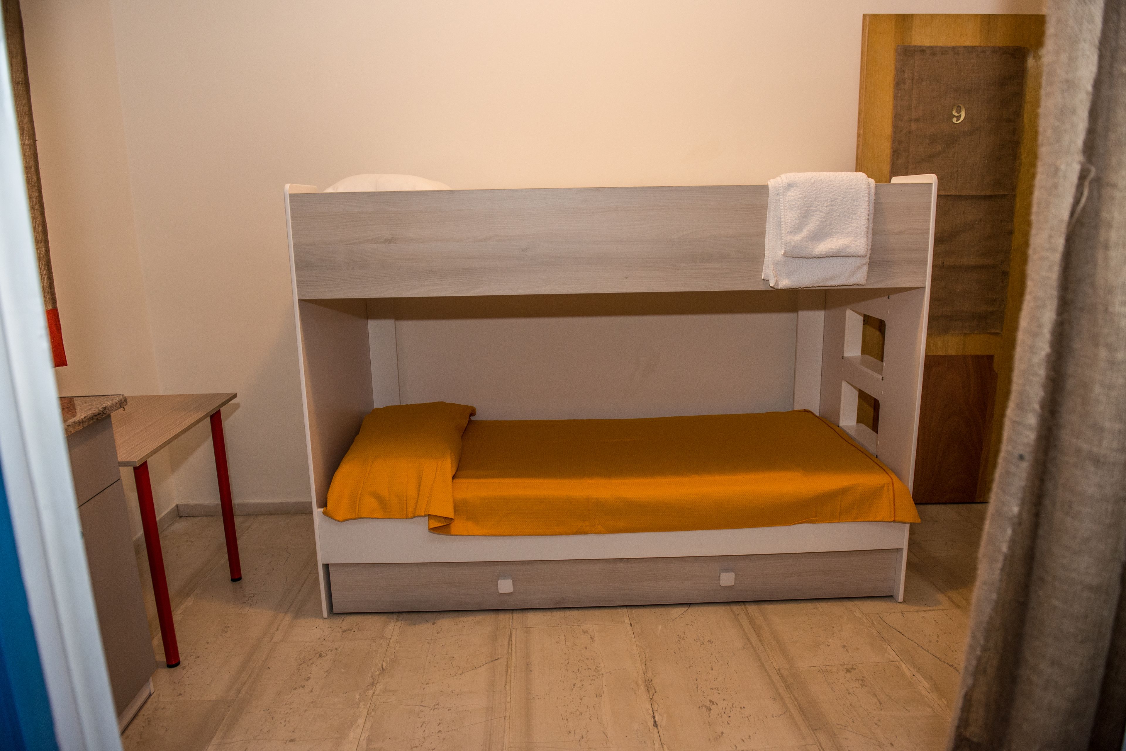 A bunk bed with a wooden headboard, a single bed below, and a small desk with a red chair.