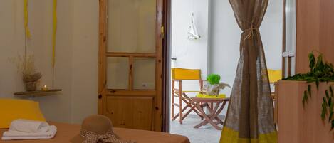 A bedroom with a bed, a wooden door, a small table with a chair, and a curtain.