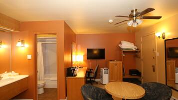 Une chambre d’hôtel avec salle de bain, un bureau équipé d’un ordinateur, une chaise, une télévision et un ventilateur de plafond.