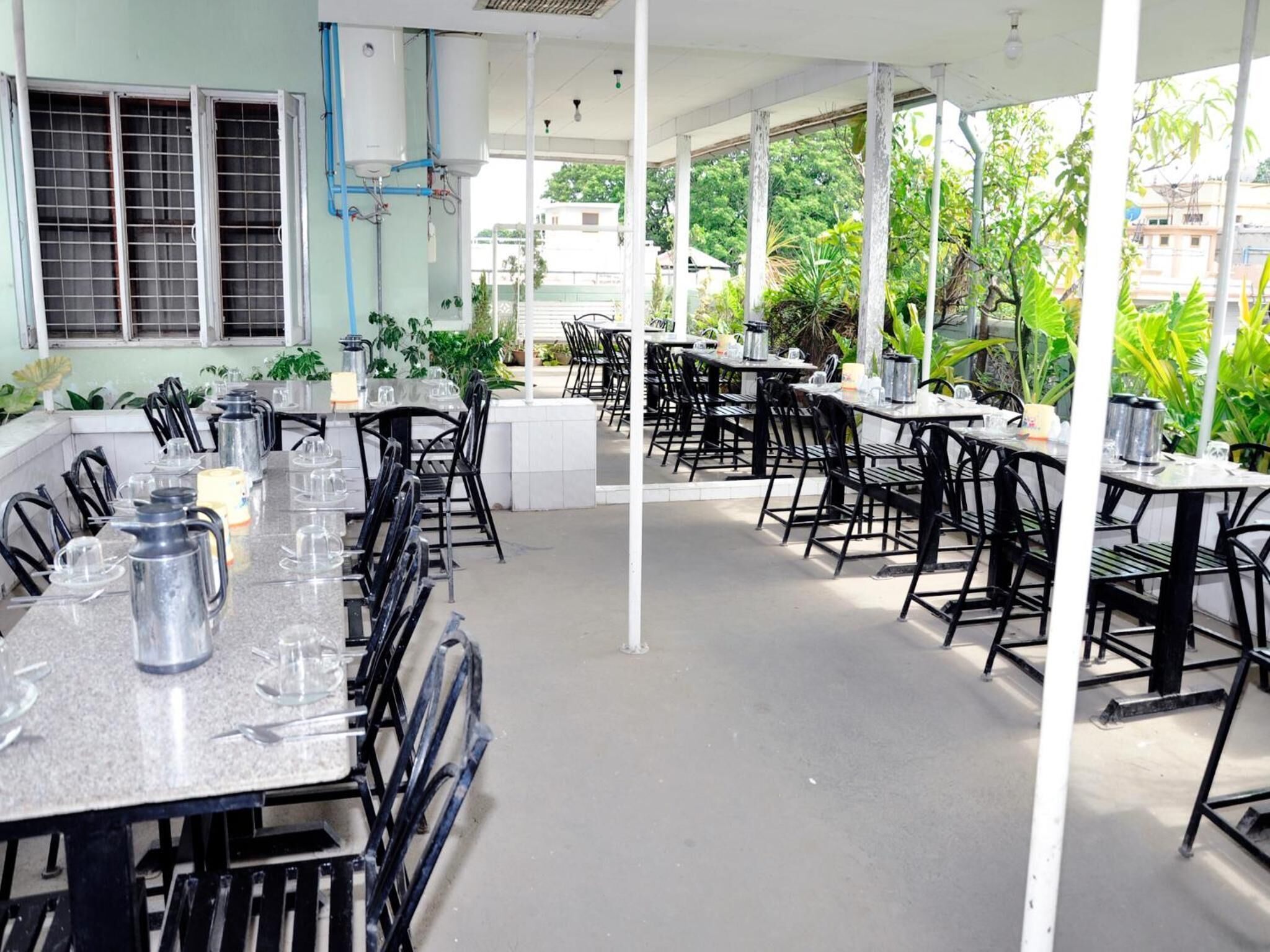An outdoor dining area with multiple tables and chairs, surrounded by greenery and buildings in the background.