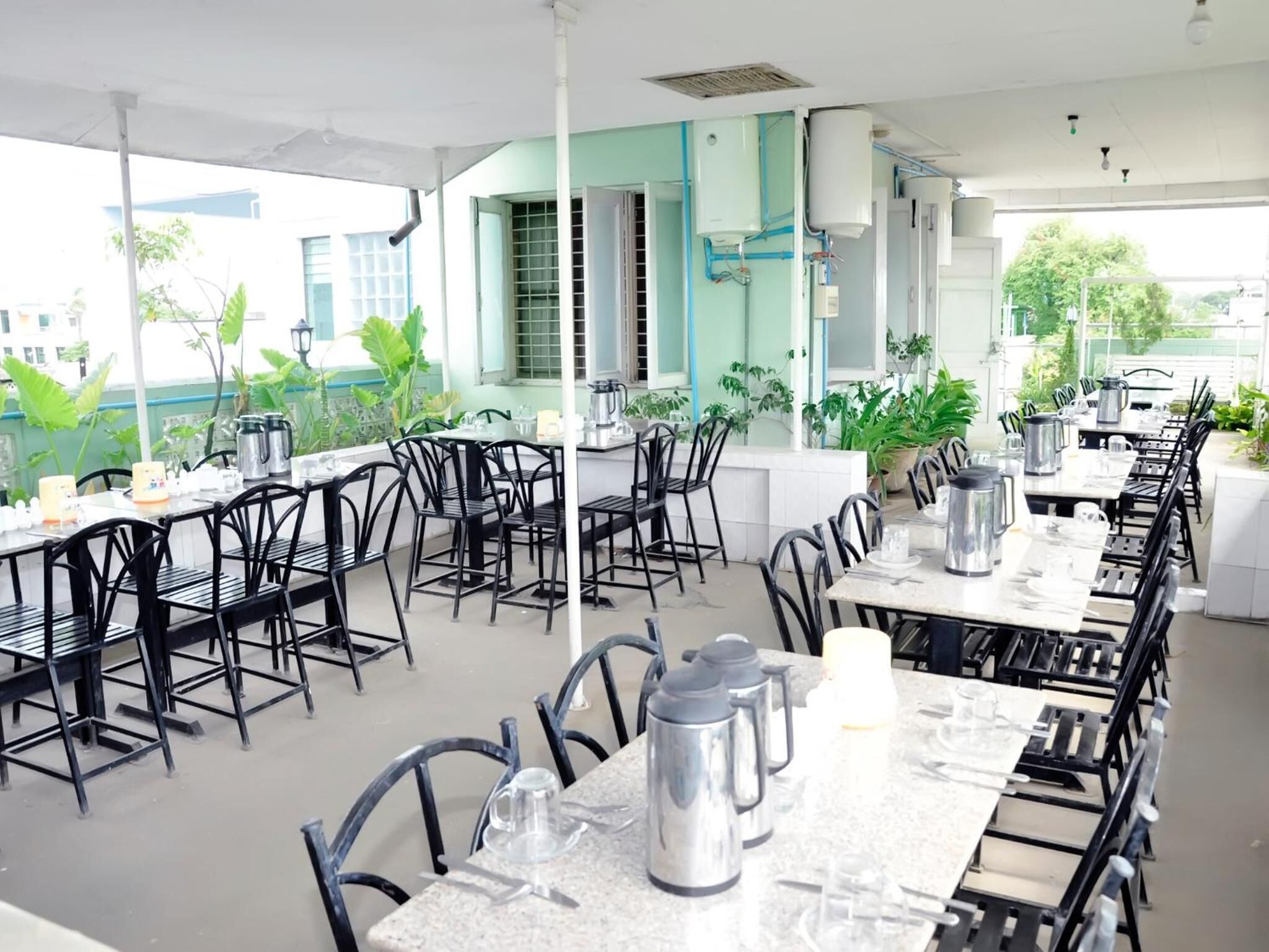 An outdoor dining area with tables and chairs, surrounded by potted plants and a building in the background.