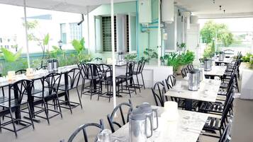 An outdoor dining area with tables and chairs, surrounded by potted plants and a building in the background.