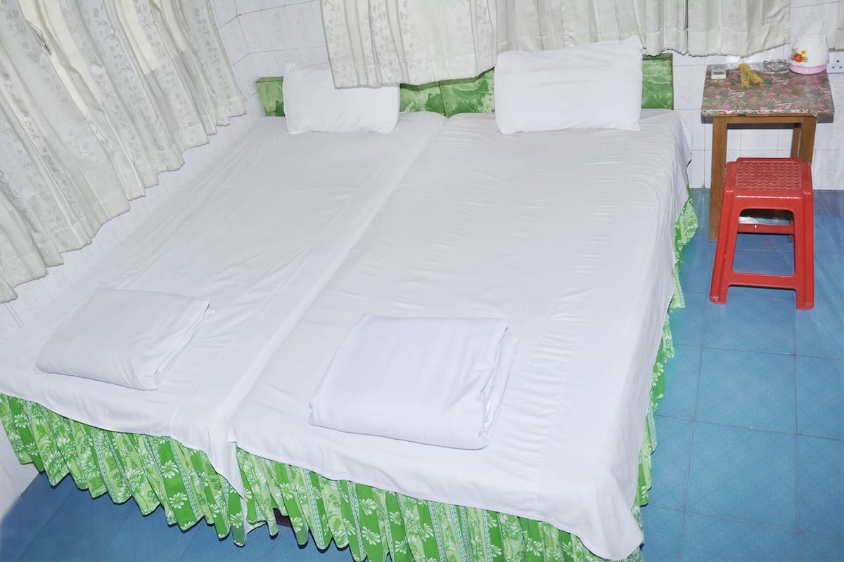 A single bed with white bedding and a green floral skirt, a small table with a floral tablecloth, and a red stool.