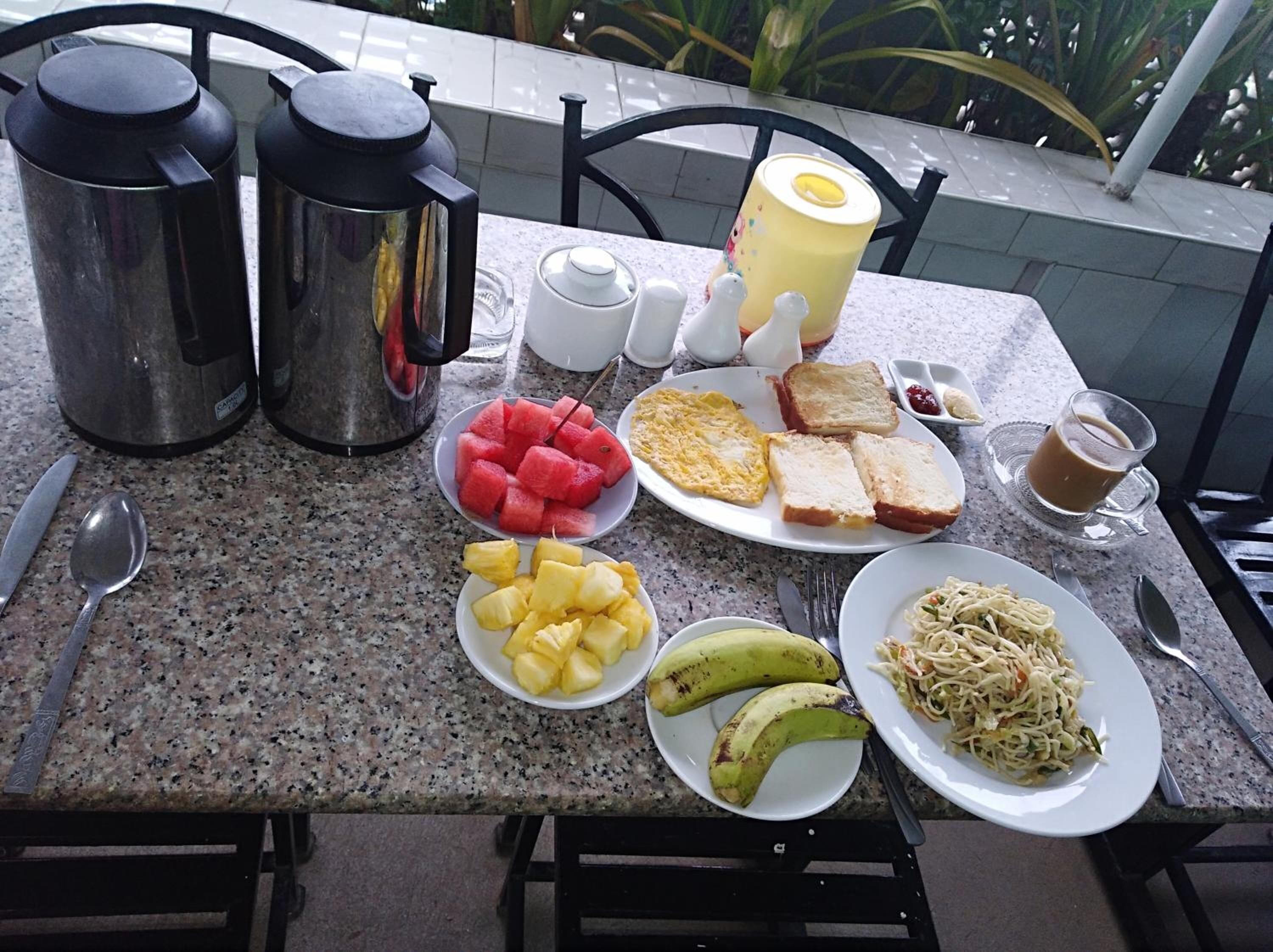Una mesa de desayuno con diversos alimentos, como tostadas, huevos, sandía, piña, plátanos y pasta, además de café y té.