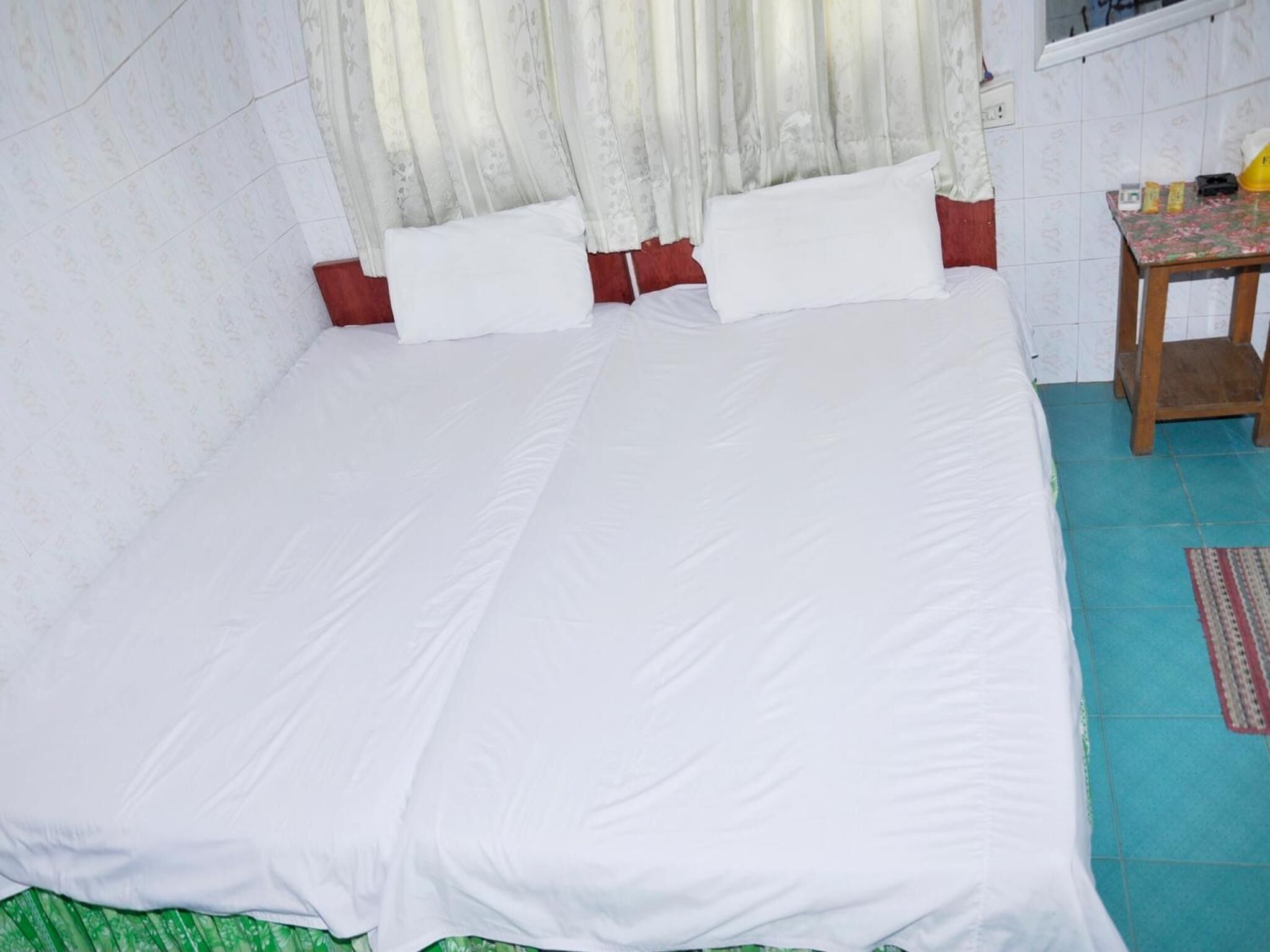A single bed with white bedding, a wooden nightstand with a lamp, and a window with curtains.