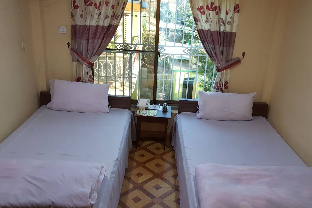 A room with two single beds, a small table with a lamp, and a window with curtains.