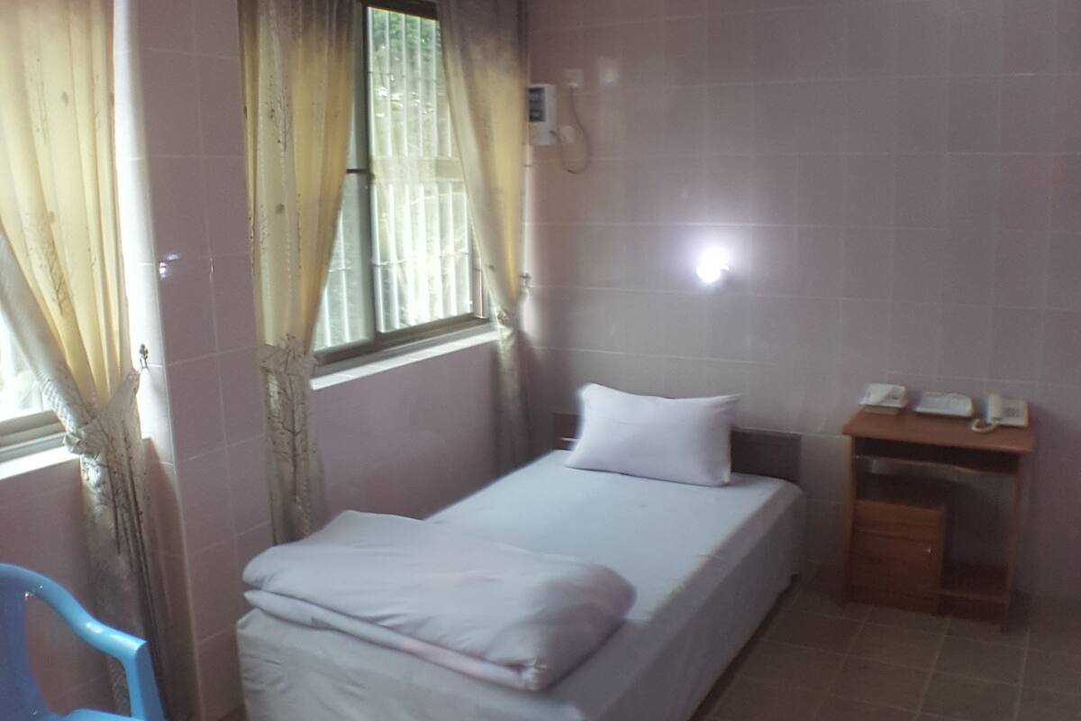 A single-bed room with a window, curtains, a small table with a telephone, and a chair.