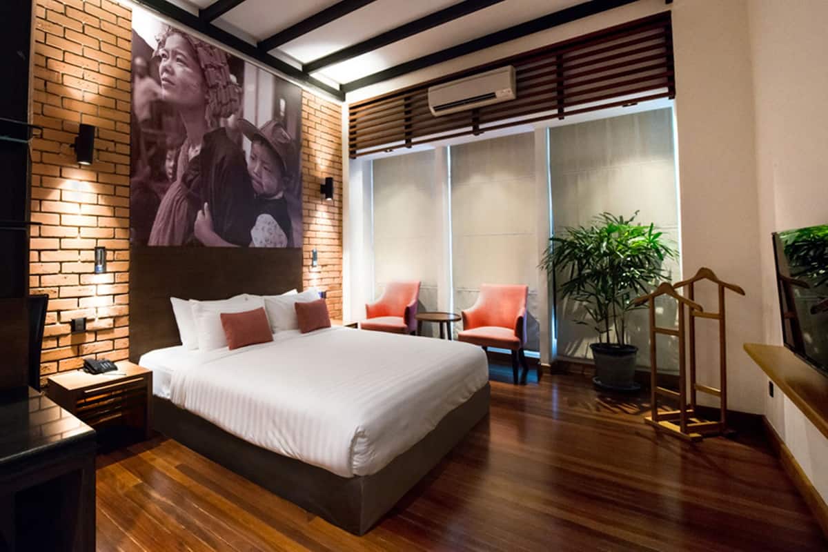 A modern hotel room with a large bed, a brick accent wall, a large window, a potted plant, and a small table with two chairs.