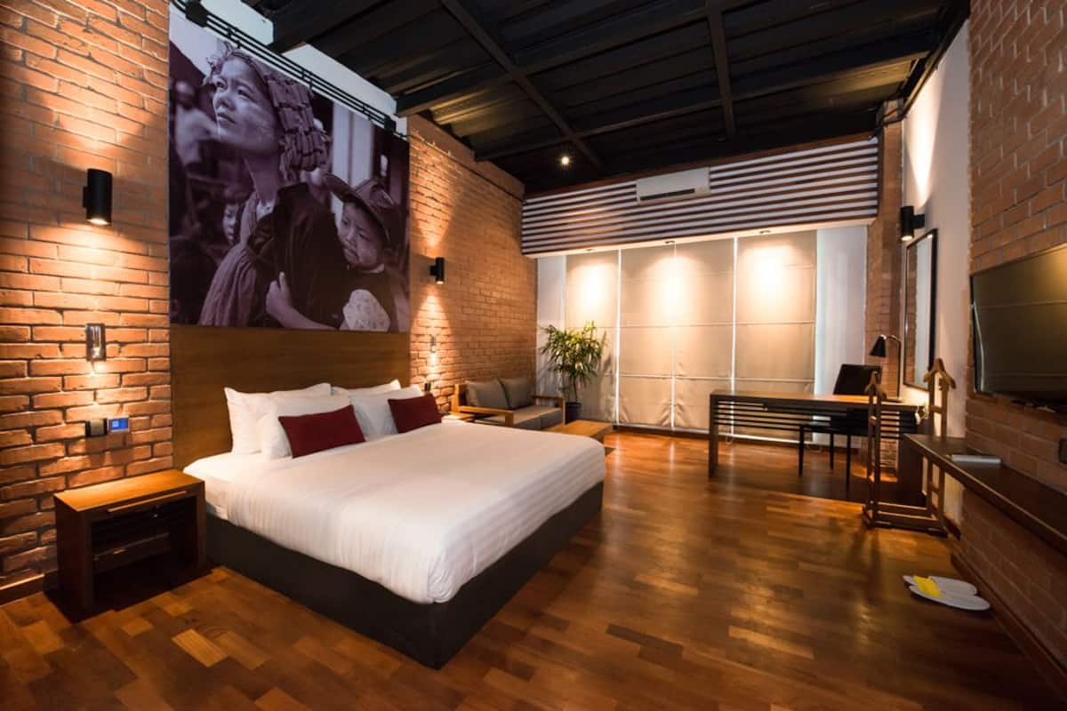 A modern hotel room with a large bed, a brick accent wall, a large window, and a flat-screen TV.